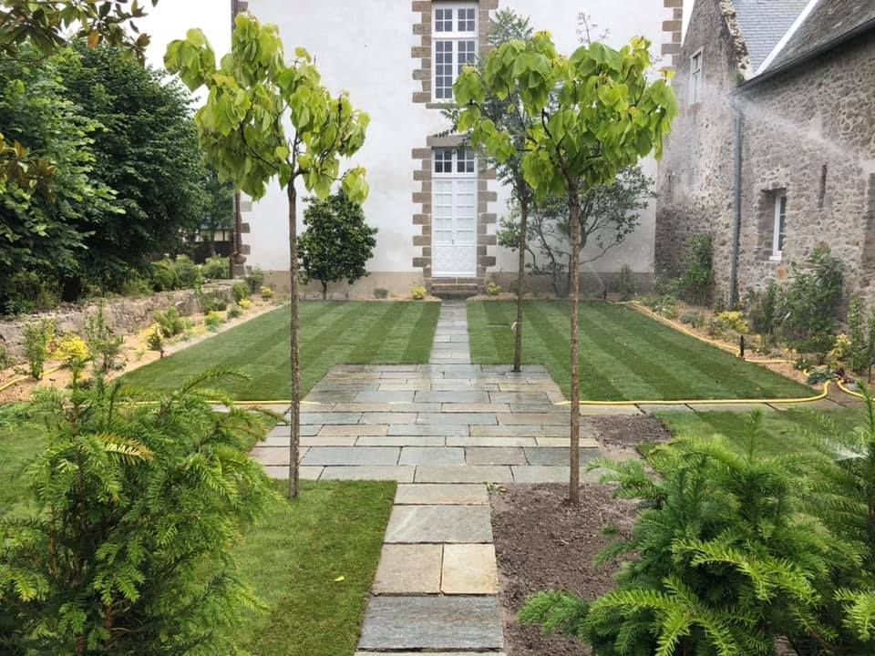 Un chemin de pierre traverse un jardin soigné et mène à une porte blanche, bordée de jeunes arbres et de haies.