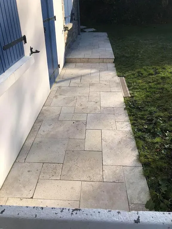 Allée en pierre longeant une maison bleue, traversant une cour herbeuse en plein jour.