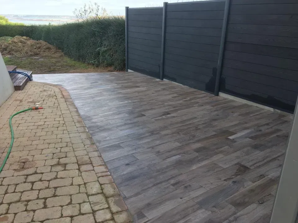 Terrasse arrière avec plancher en bois gris, bordée d'un chemin de briques et d'une clôture sombre assurant l'intimité.