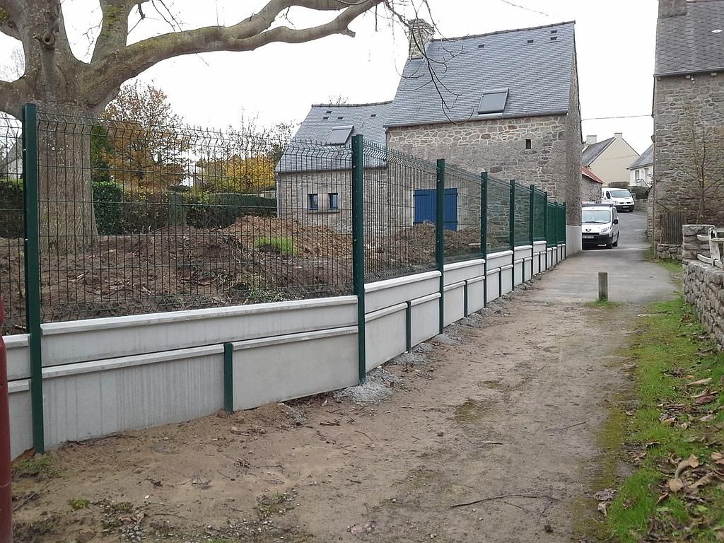 Un étroit chemin de gravier longe une route clôturée, avec des maisons en pierre et une voiture garée au loin.