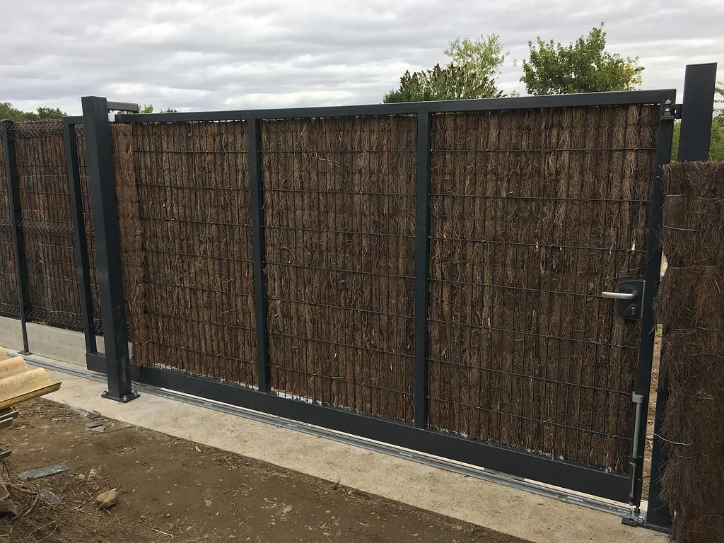 Clôture en métal noir avec panneaux d'intimité tissés le long d'une allée de terre sous un ciel nuageux.