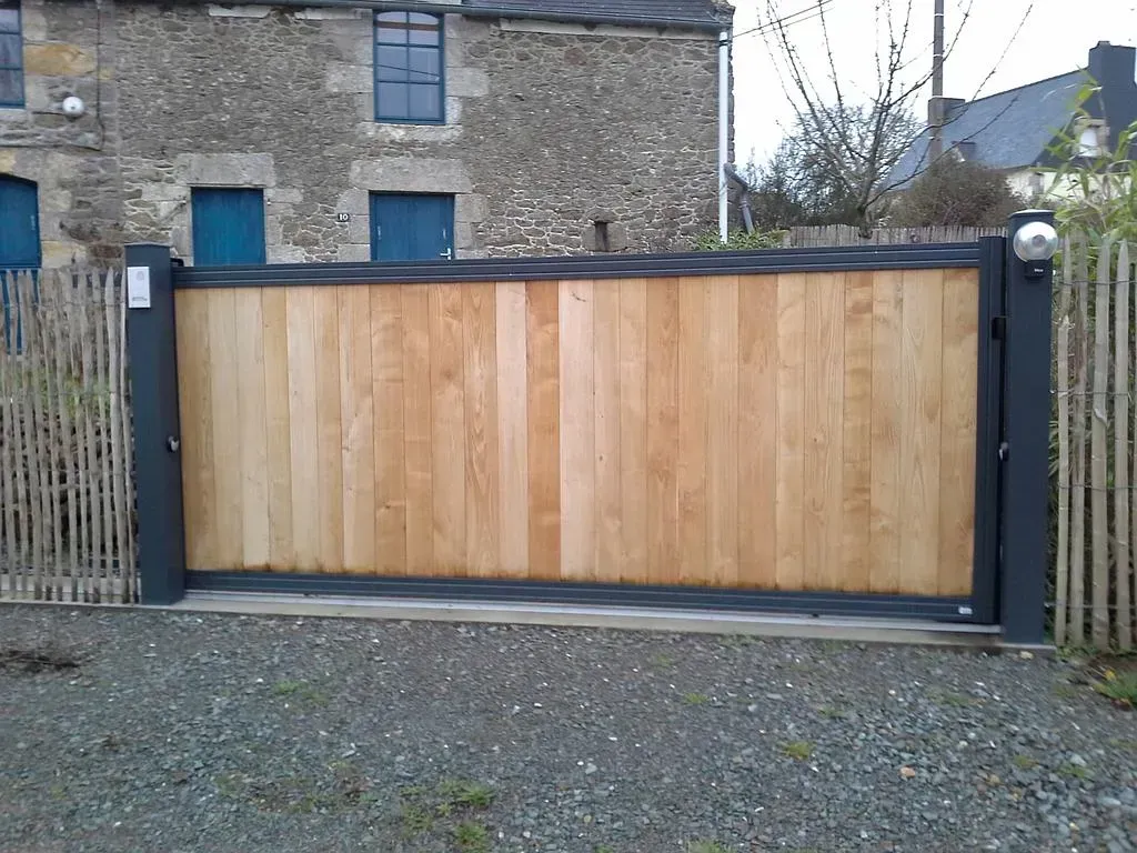 Portail coulissant en bois avec poteaux noirs devant une maison en pierre.