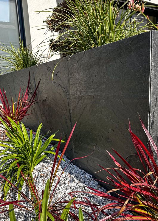 Mur moderne en pierre sombre avec des plantes ornementales rouges et vertes le long d'un lit de gravier.