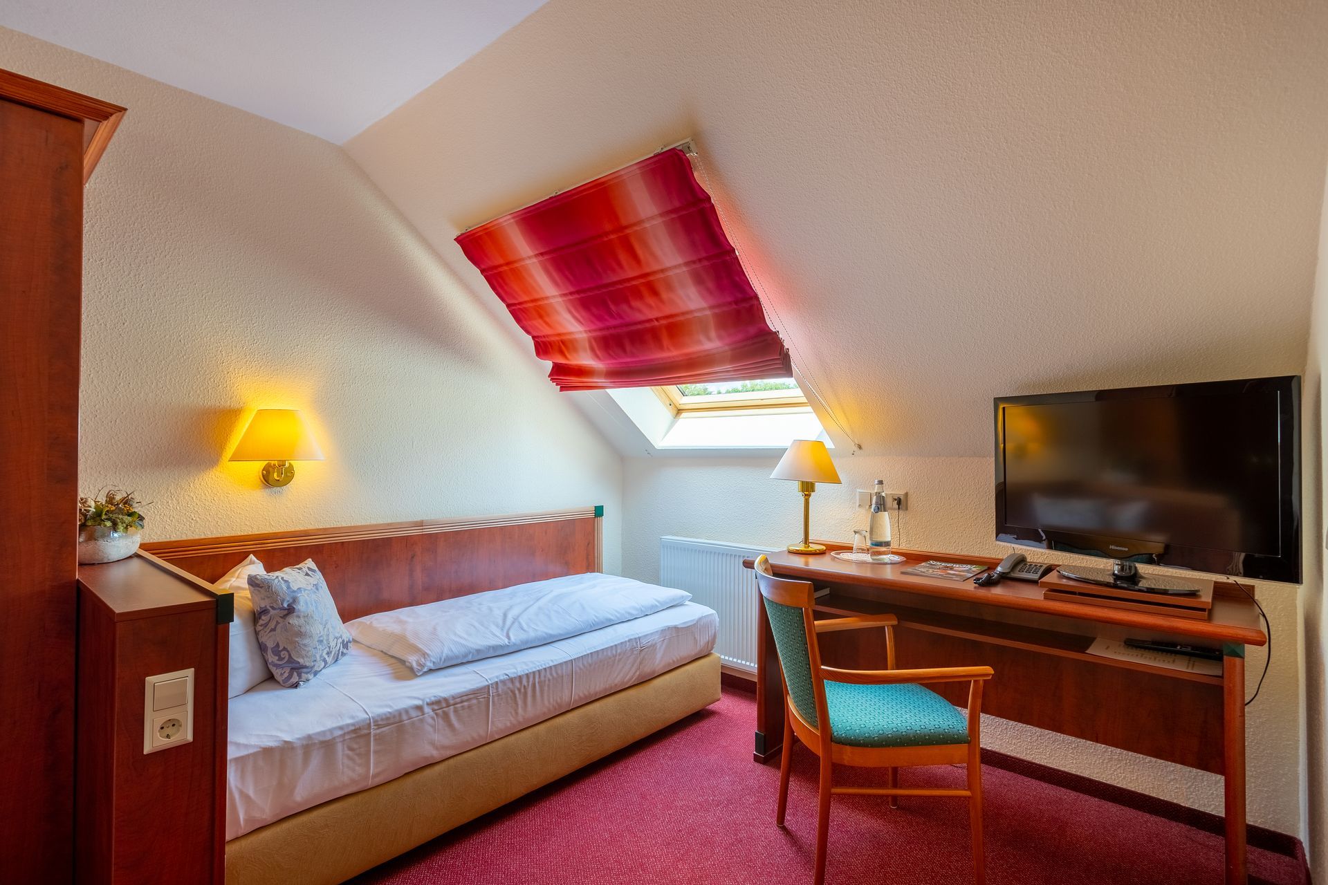 Kleines Hotelzimmer mit zwei Einzelbetten, rotem Teppich, schrägem Dachfenster, Schreibtisch und Fernseher