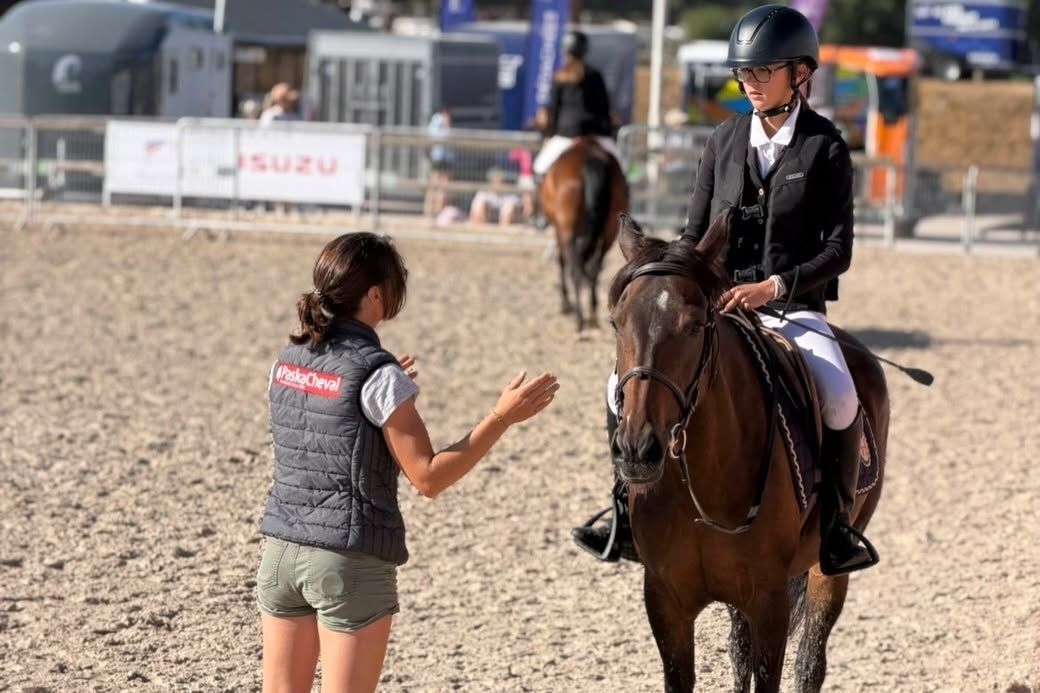 Une femme parle à un cavalier à cheval dans un manège en plein air, avec un autre cheval à l’arrière-plan