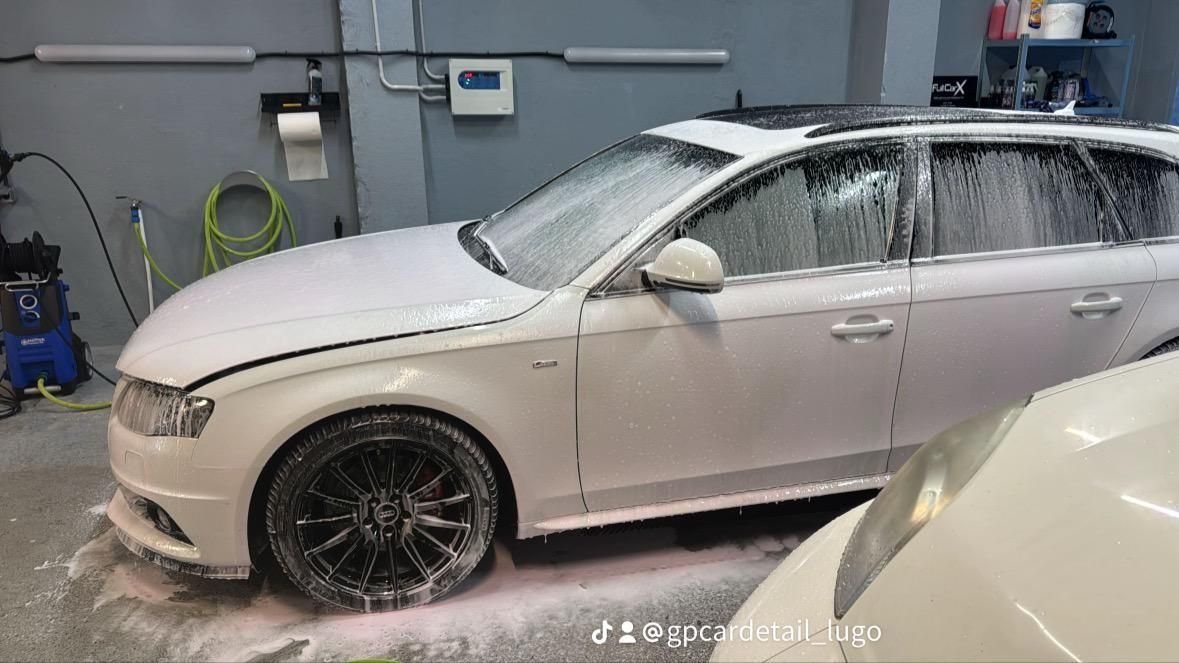 SUV blanco cubierto de espuma en una cabina de lavado de coches, con un segundo coche parcialmente visible en primer plano.