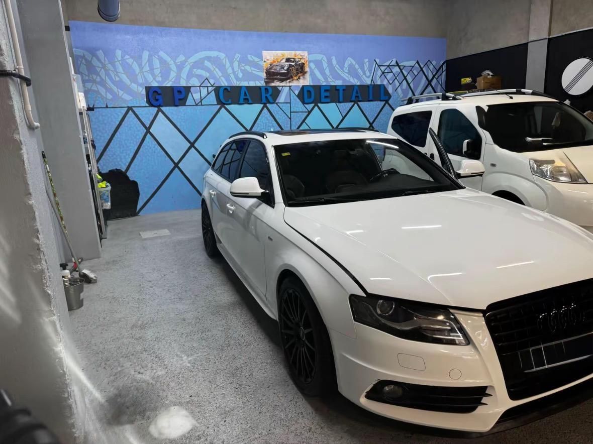 Un sedán blanco estacionado en un garaje junto a otro coche, con un mural azul en la pared.