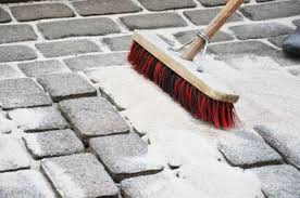 Eine Person fegt mit einem rotborstigen Besen weißen Sand in die Fugen grauer Pflastersteine.