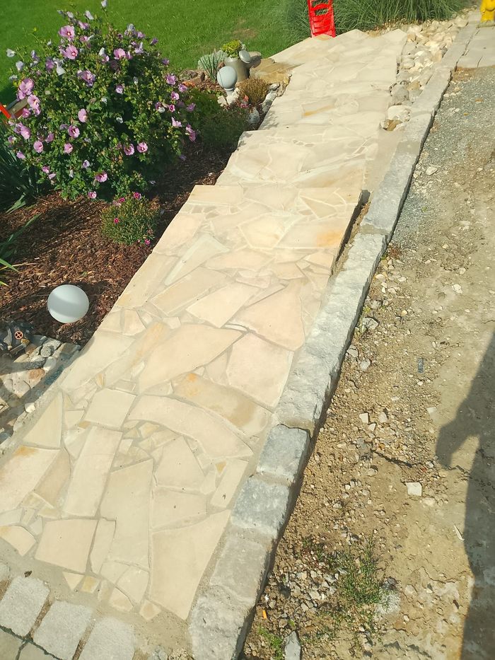 Steinweg mit unebenen Steinplatten, eingefasst mit grauen Ziegeln, neben Garten und Kies.