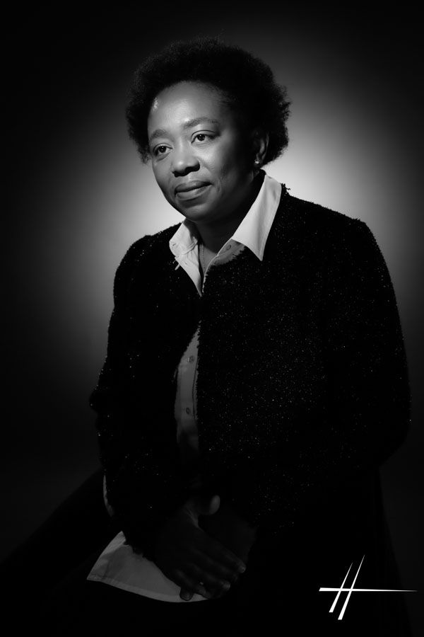 Portrait en studio, en noir et blanc, d'une femme assise portant une veste sombre, doucement éclairée sur un fond sombre.