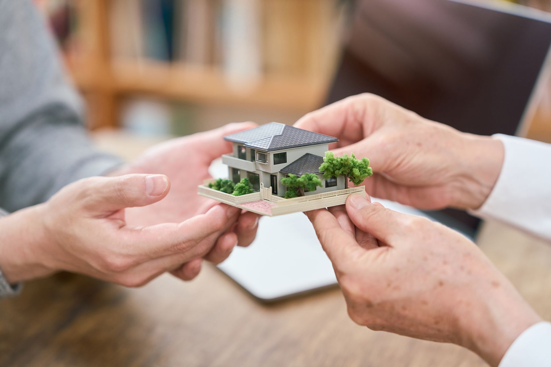 Des mains tenant une petite maquette de maison au-dessus d’une table en bois, avec un ordinateur portable en arrière-plan.