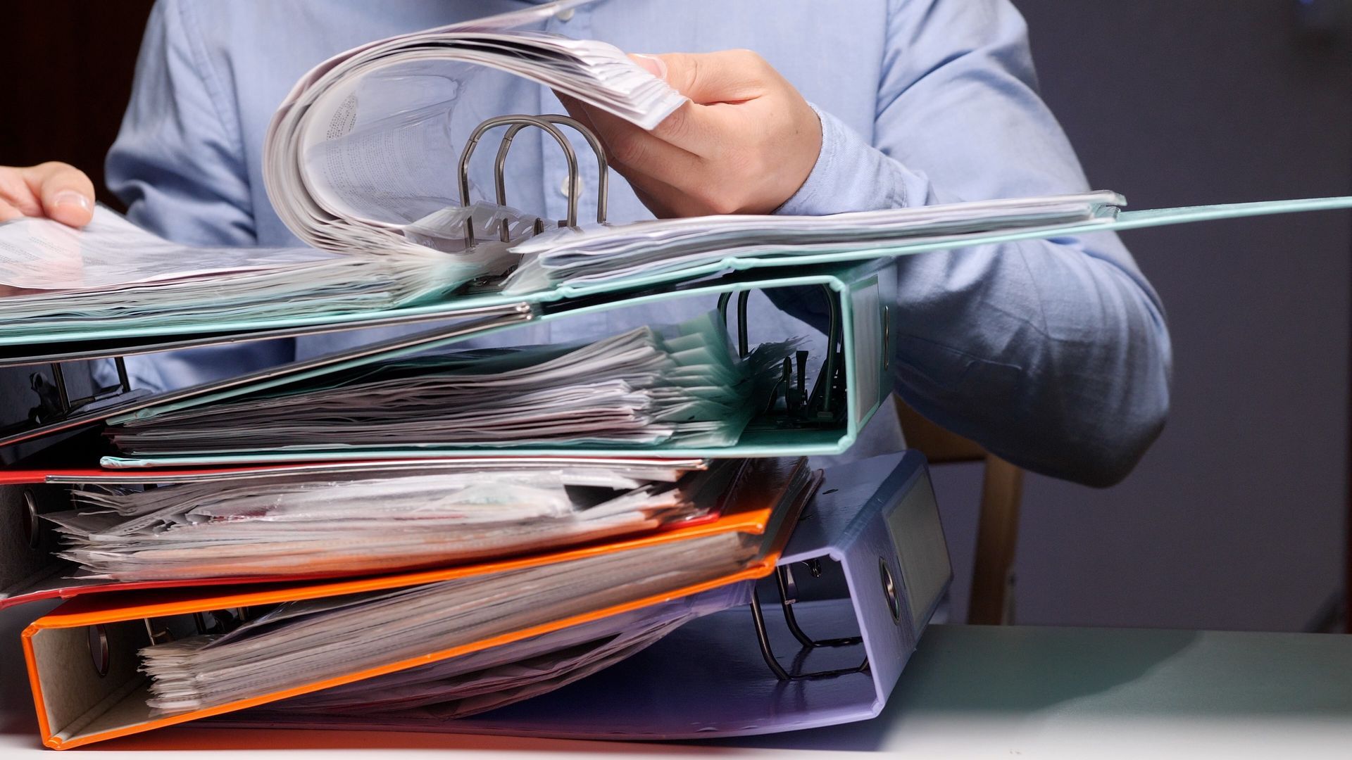 Des mains fouillent dans une pile de dossiers et de chemises débordant sur un bureau