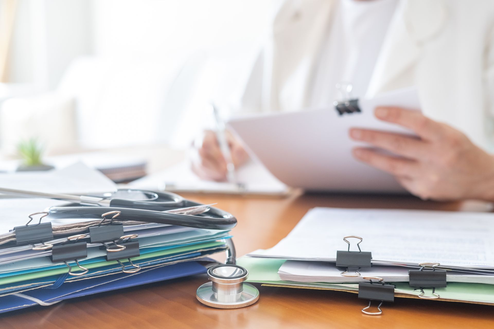 Un professionnel de santé prend des notes sur un bloc-notes à côté d'un stéthoscope et de dossiers médicaux empilés sur un bureau.