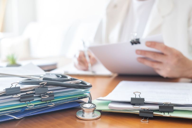Un professionnel de santé prend des notes sur un bloc-notes à côté d'un stéthoscope et de dossiers médicaux empilés sur un bureau.