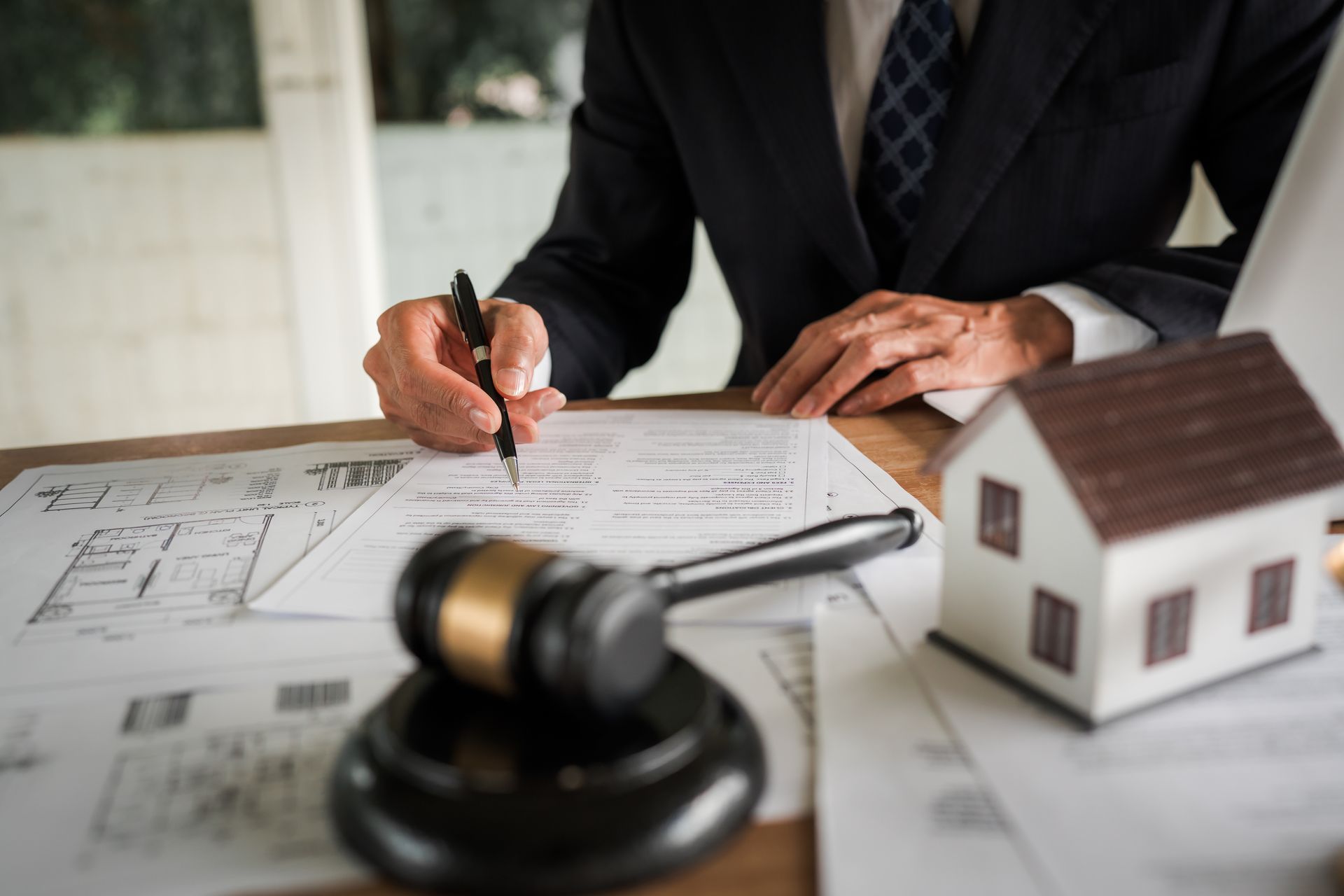 Un avocat signe des documents immobiliers à côté d’une maquette de maison et d’un marteau de juge posés sur un bureau