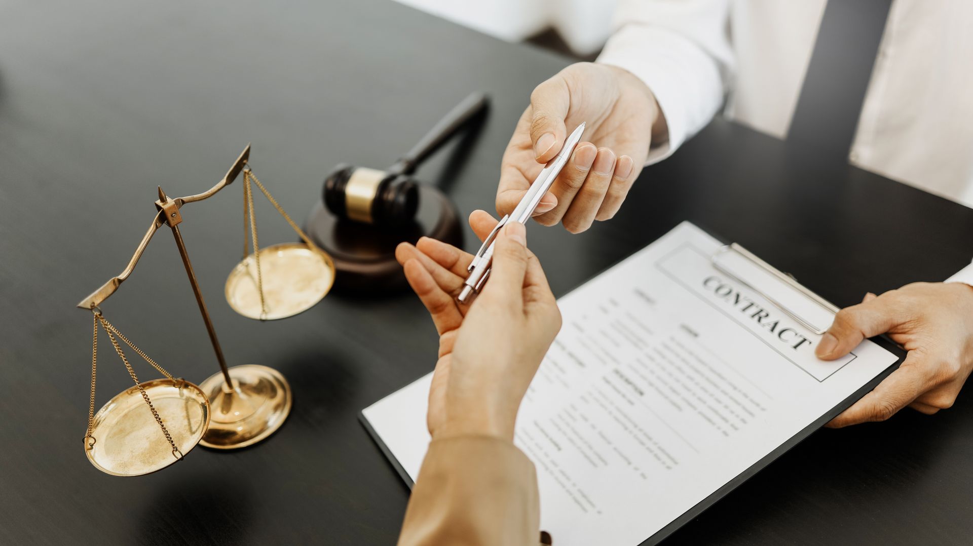 Des mains se tendant un stylo au-dessus d’un contrat, à côté d’une balance en laiton et d’un maillet de juge posés sur un bureau