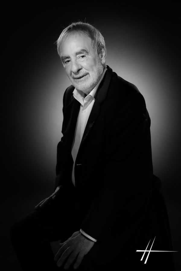Portrait en noir et blanc d'un homme assis en costume, souriant doucement sur un fond sombre.