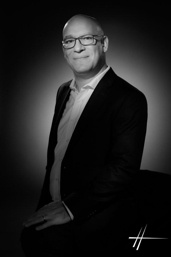 Portrait en studio d'un homme chauve portant des lunettes et un costume sombre, sur fond sombre.