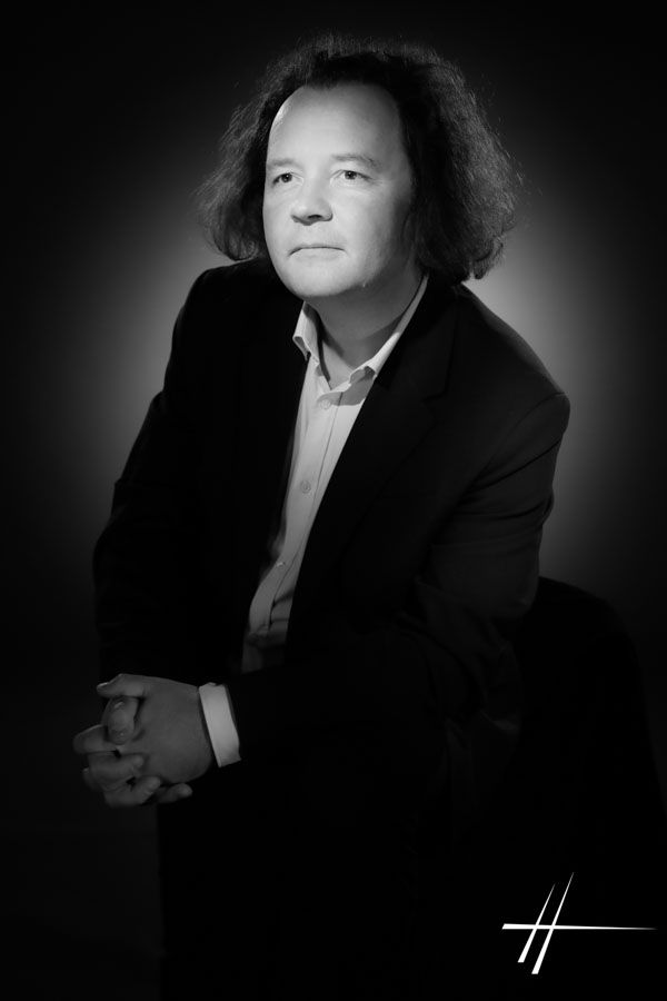 Portrait en noir et blanc d'une personne en costume sombre, assise sur fond sombre, avec une signature dans le coin.