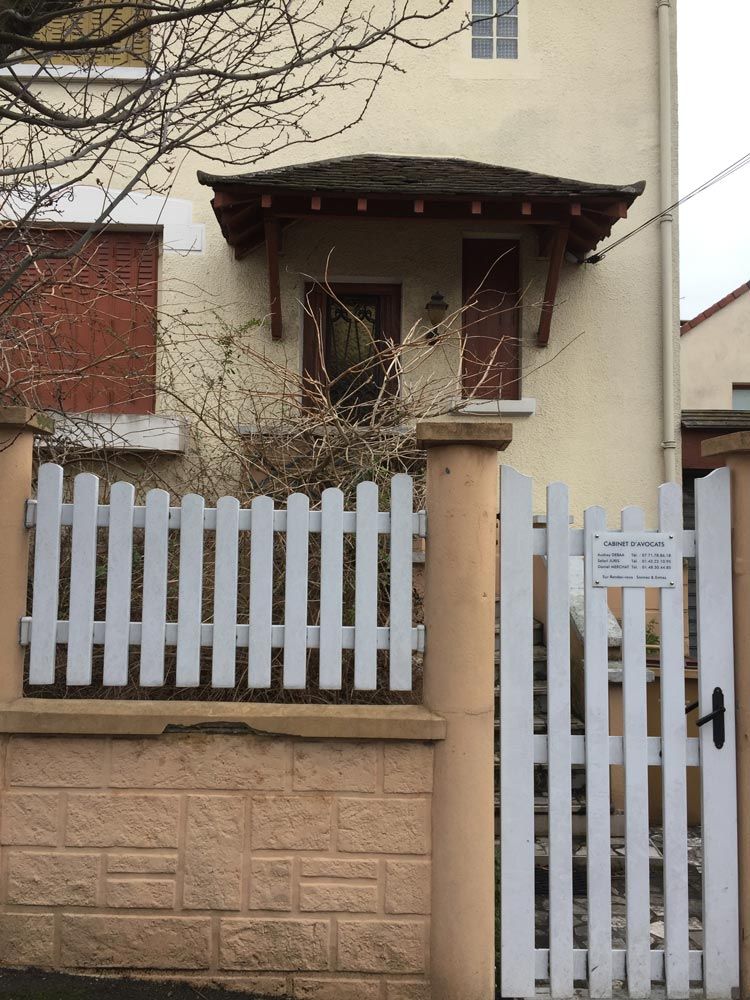Petite maison avec un porche, une clôture blanche et un portail fermé derrière un mur de briques.