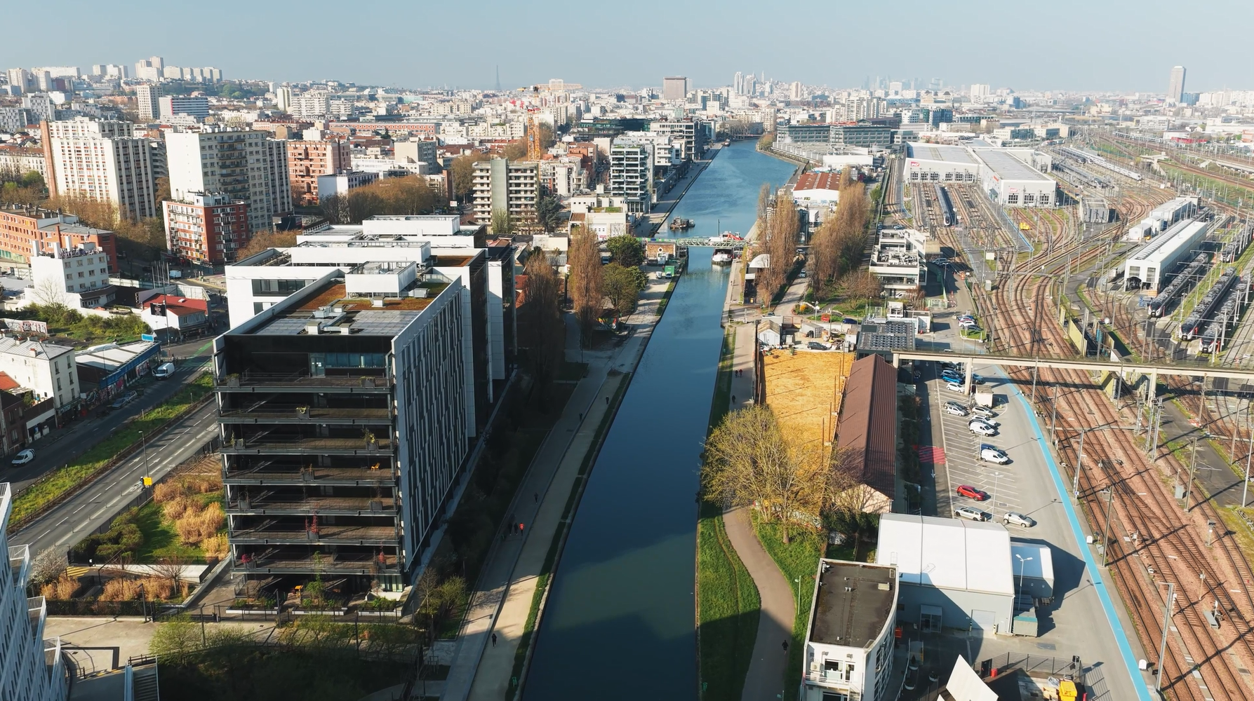 Vue aérienne d'un canal traversant une ville dense, avec des bâtiments et des voies ferrées de part et d'autre.