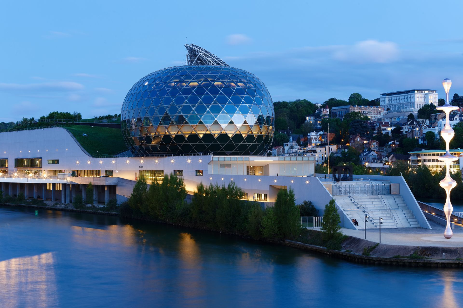 Bâtiment en verre en forme de dôme illuminé au crépuscule au bord d'une rivière, avec une promenade moderne en bord de rivière et des escaliers.