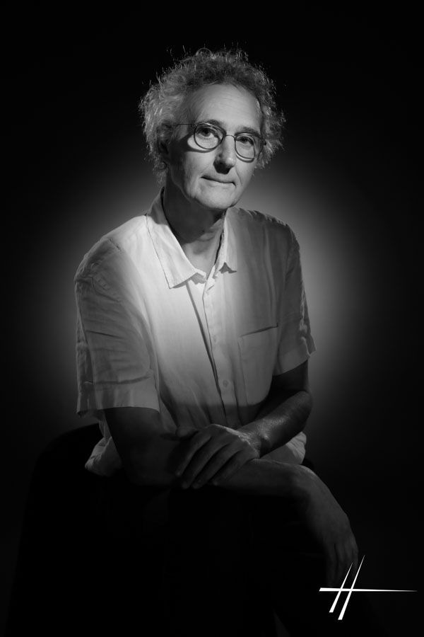 Portrait en noir et blanc d'une personne assise, vêtue d'une chemise claire, doucement éclairée sur un fond sombre.