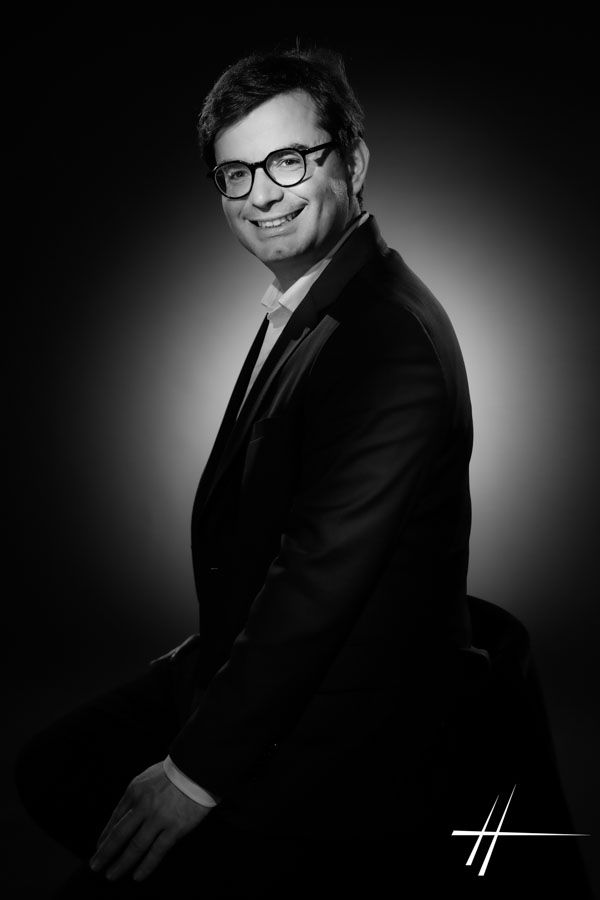 Un homme souriant, vêtu d'un costume sombre et portant des lunettes, pose devant un fond noir.