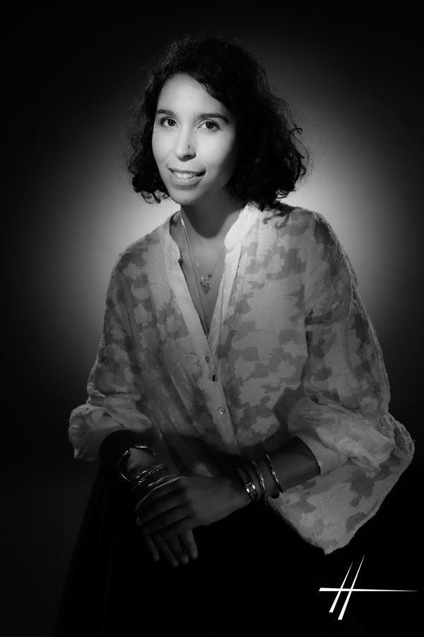 Portrait en noir et blanc, réalisé en studio, d'une personne assise portant un chemisier à motifs, légèrement éclairée sur fond sombre.