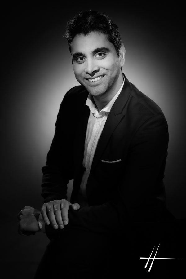 Portrait en noir et blanc, réalisé en studio, d'une personne souriante en blazer sur fond sombre.