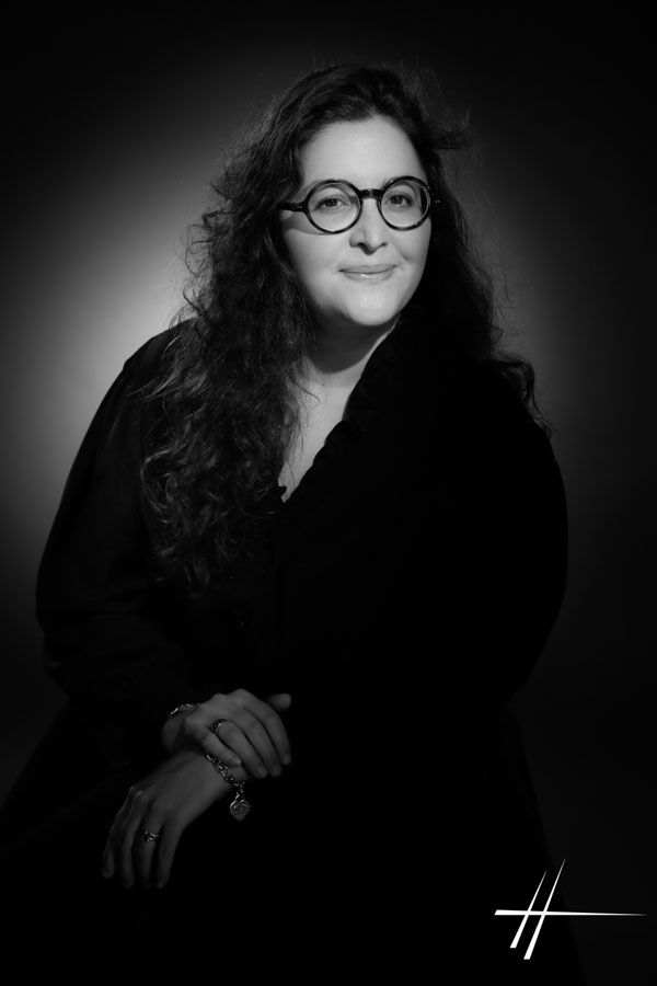 Portrait en noir et blanc d'une personne aux longs cheveux bouclés et portant des lunettes, assise sur un fond sombre.