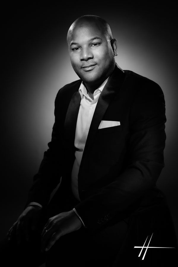 Portrait en studio d'un homme en costume sombre, assis sur fond sombre, regardant l'objectif.