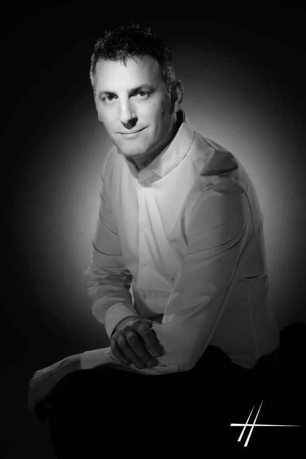 Portrait en noir et blanc d'un homme assis, penché en avant, portant une chemise claire sur fond sombre.