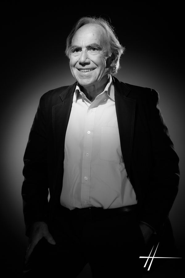 Portrait en noir et blanc d'une personne souriante portant un blazer foncé et une chemise blanche sur fond sombre.