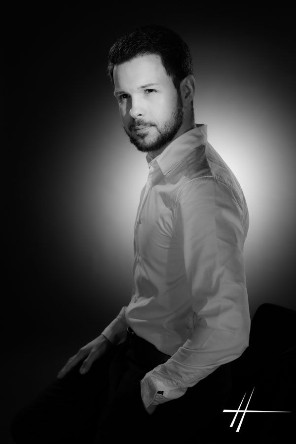 Portrait en noir et blanc, en studio, d'un homme en chemise blanche, assis et tourné vers l'objectif, éclairé par un projecteur à lumière douce.