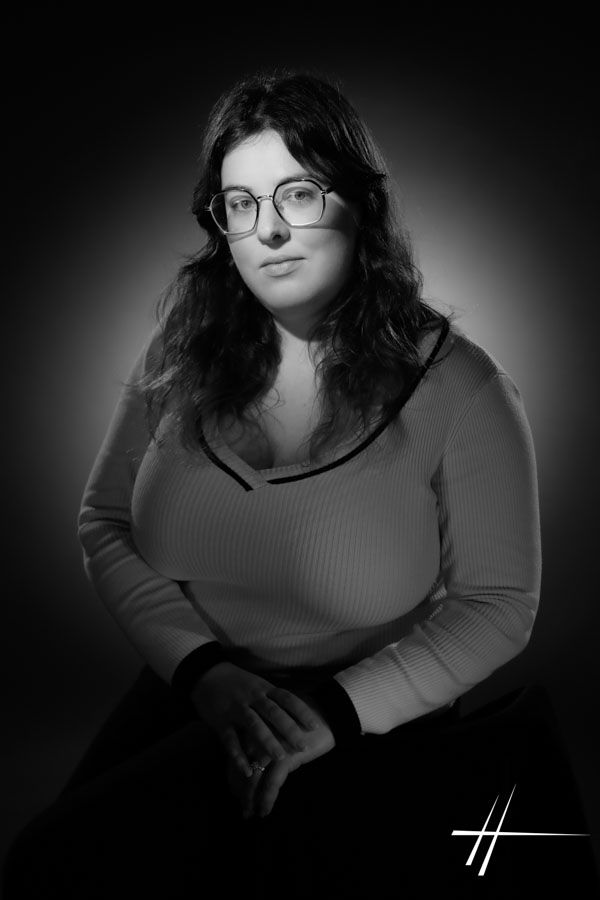 Portrait en noir et blanc d'une personne assise, portant des lunettes et des cheveux longs, face à l'objectif sur fond sombre.
