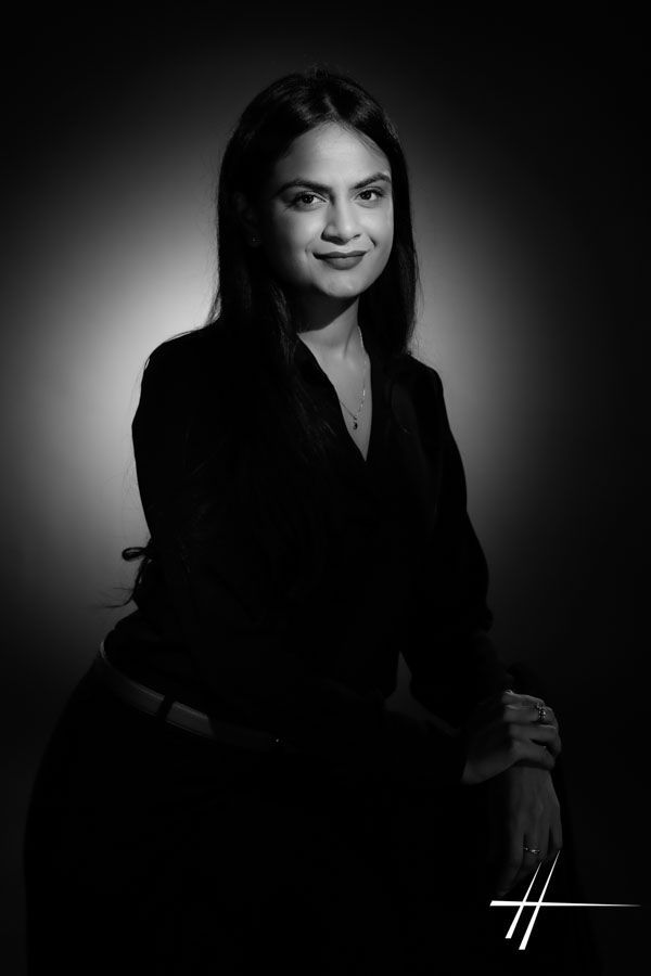 Portrait en noir et blanc d'une personne assise, vêtue de vêtements sombres, sur un fond de studio doux.