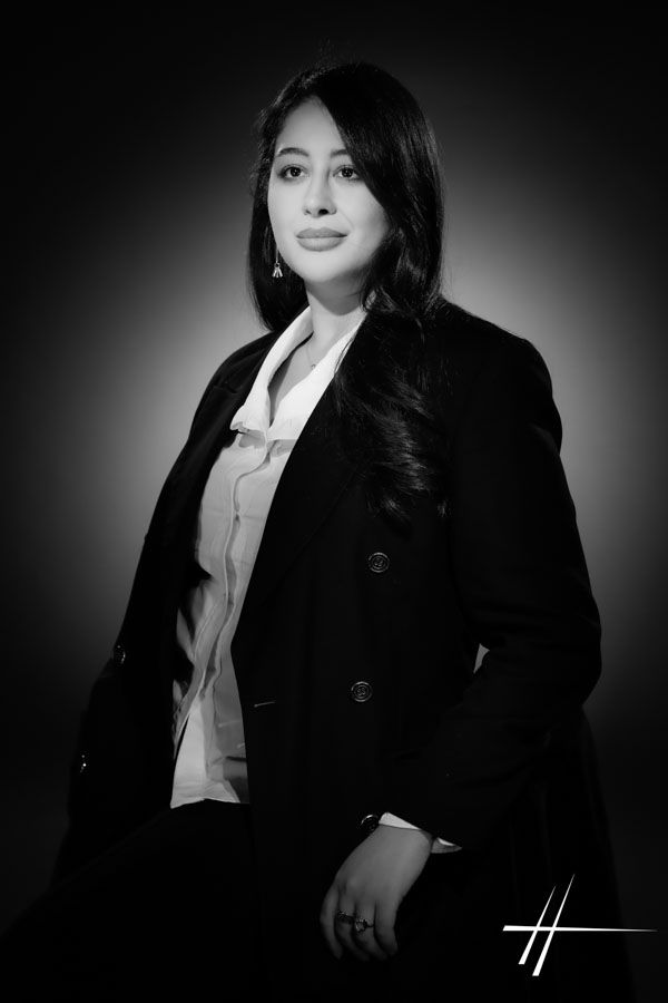 Portrait en noir et blanc, réalisé en studio, d'une personne portant un blazer foncé et un chemisier blanc sur fond sombre.