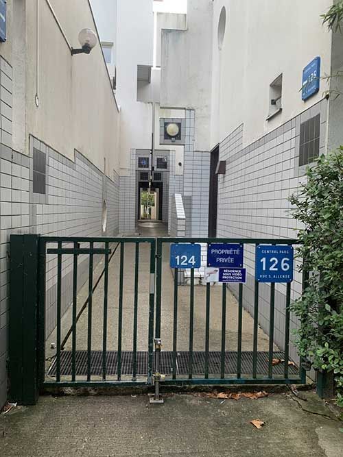 Étroit passage fermé par un portail entre des bâtiments blancs, avec les plaques d’adresse bleues.