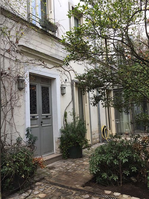 Cour étroite avec une porte grise, des murs en pierre blanche et des plantes en pot sous un arbre feuillu.