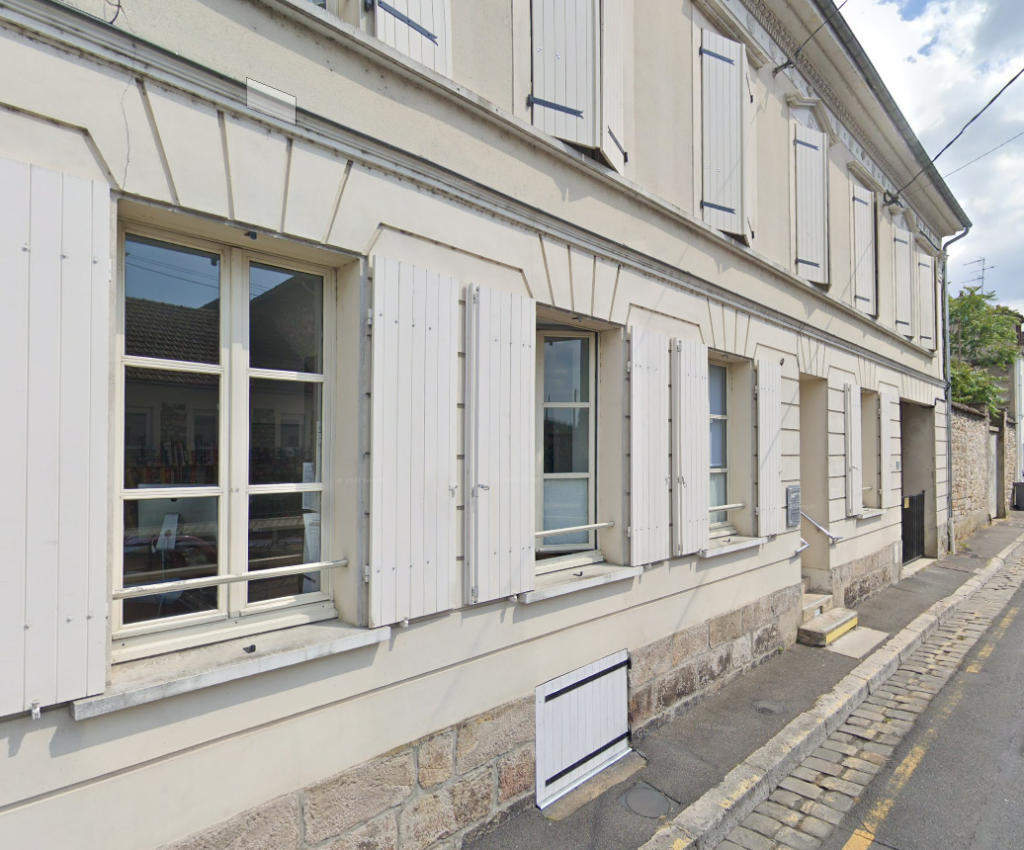 Bâtiment blanc aux fenêtres à volets clos, le long d'une rue pavée étroite par temps couvert.