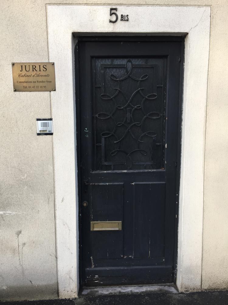 Porte d'entrée noire ornée de ferronneries décoratives, encastrée dans un mur beige.