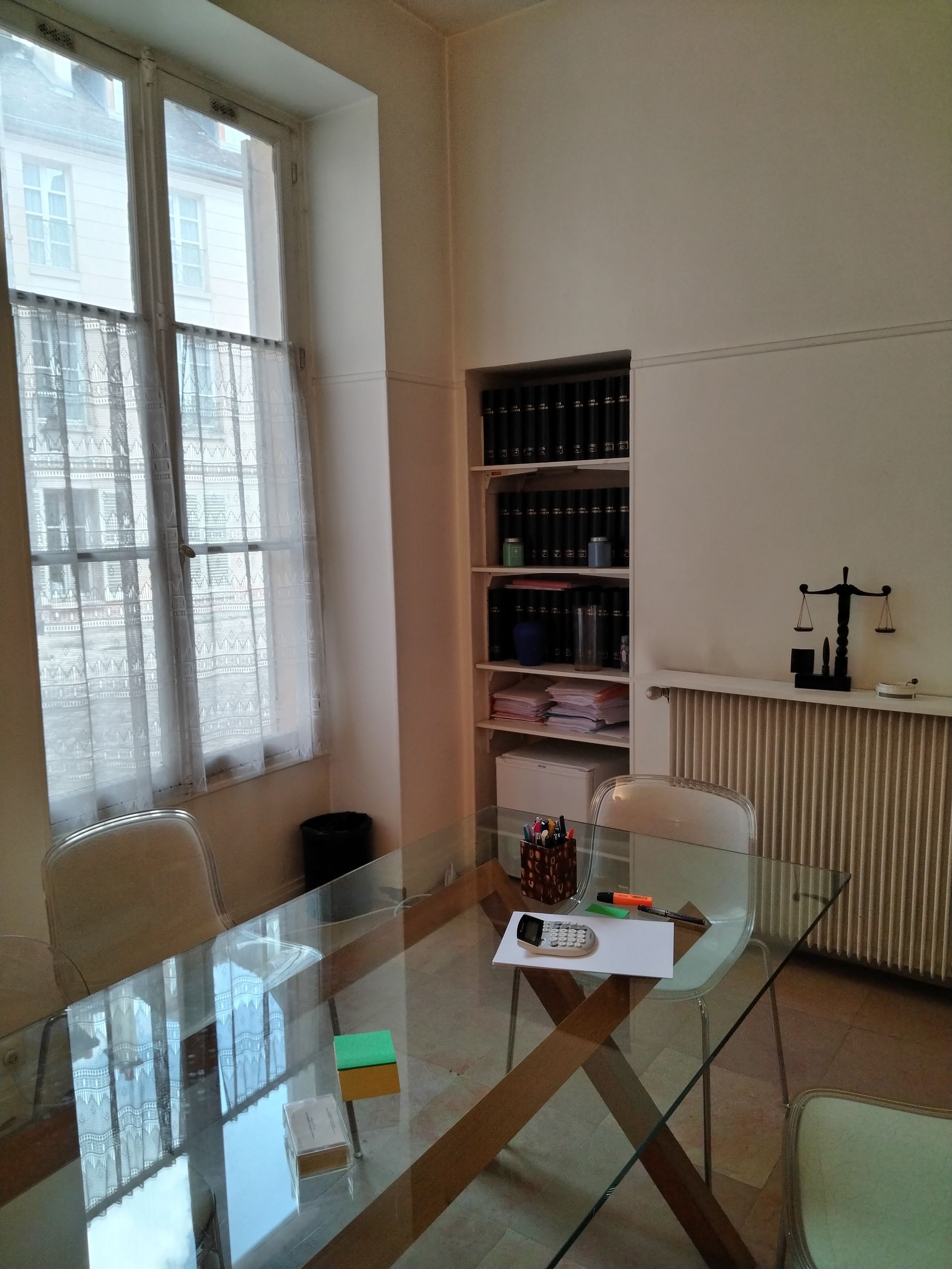 Pièce lumineuse avec un bureau en verre, une bibliothèque, une grande fenêtre avec rideaux.