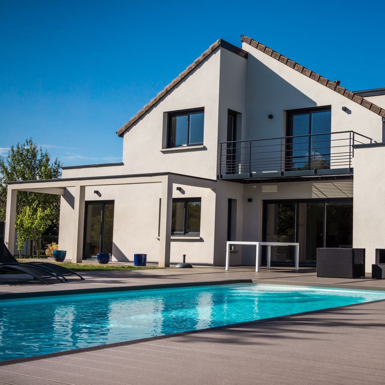 Une maison moderne beige de deux étages avec un balcon donnant sur une piscine rectangulaire.