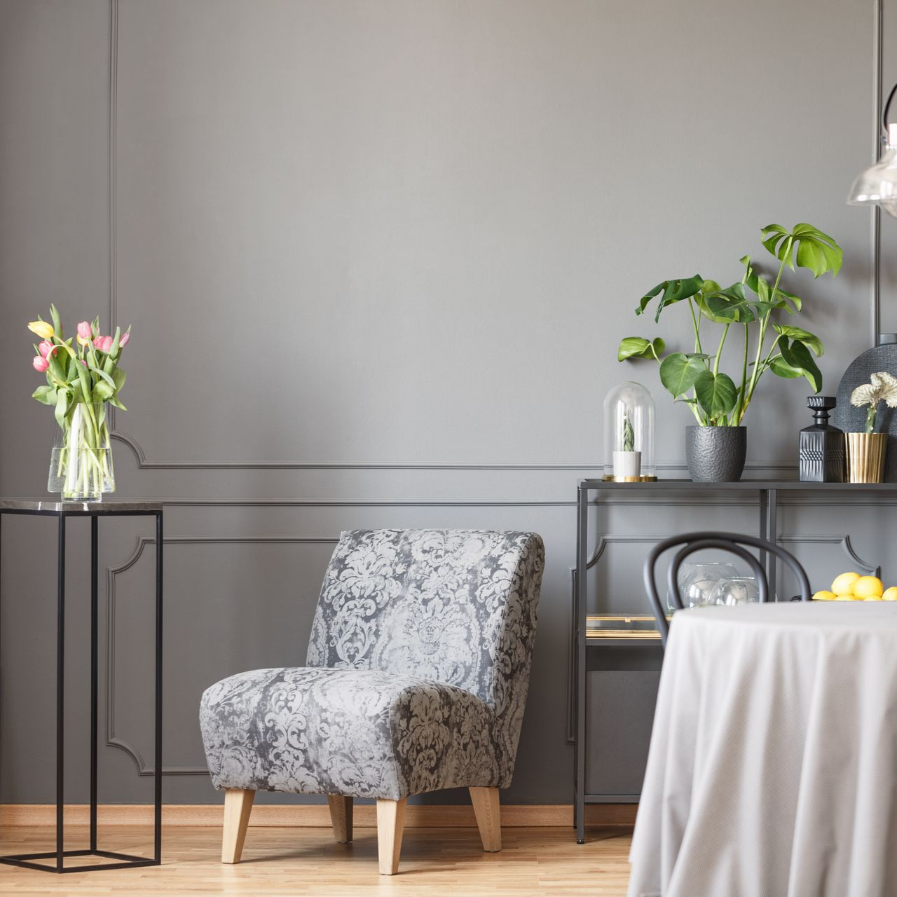 Une pièce aux murs gris présente un fauteuil à motifs floraux, un vase en verre rempli de tulipes sur un piédestal.