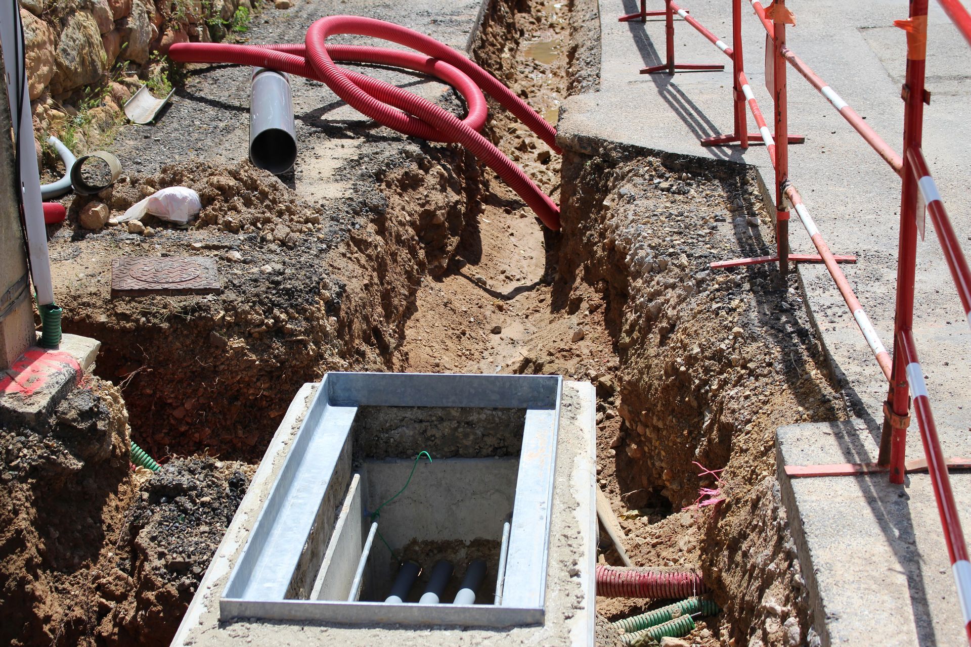 Une tranchée de chantier avec un regard de drainage en béton, des tuyaux rouges et des barrières de sécurité sur un trottoir.