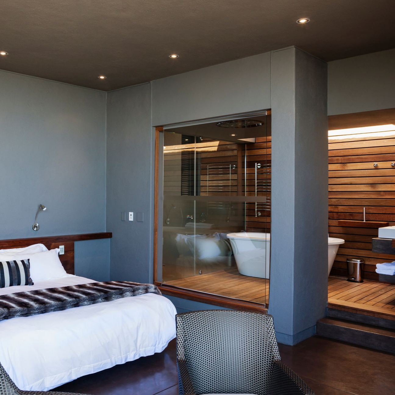 Une chambre d'hôtel moderne comprenant un lit, un fauteuil gris et une salle de bains ouverte attenante avec baignoire en îlot.