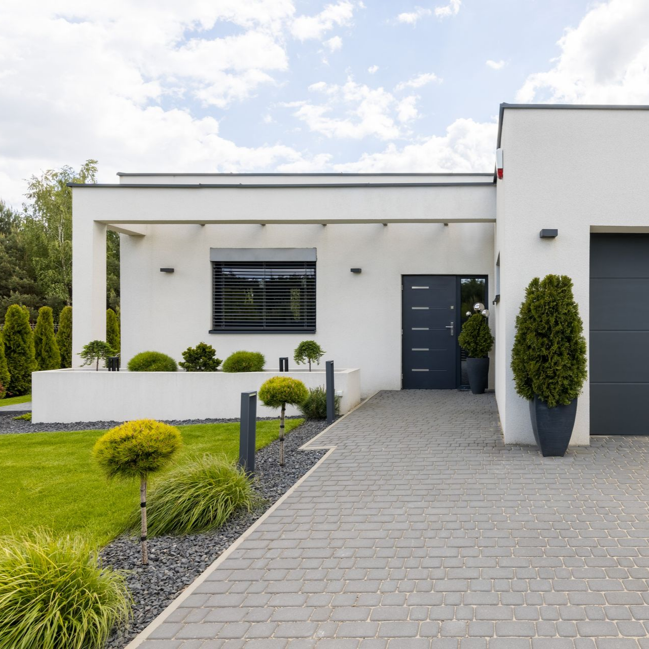 Maison blanche moderne avec une porte grise, une allée pavée et un aménagement paysager soigné.