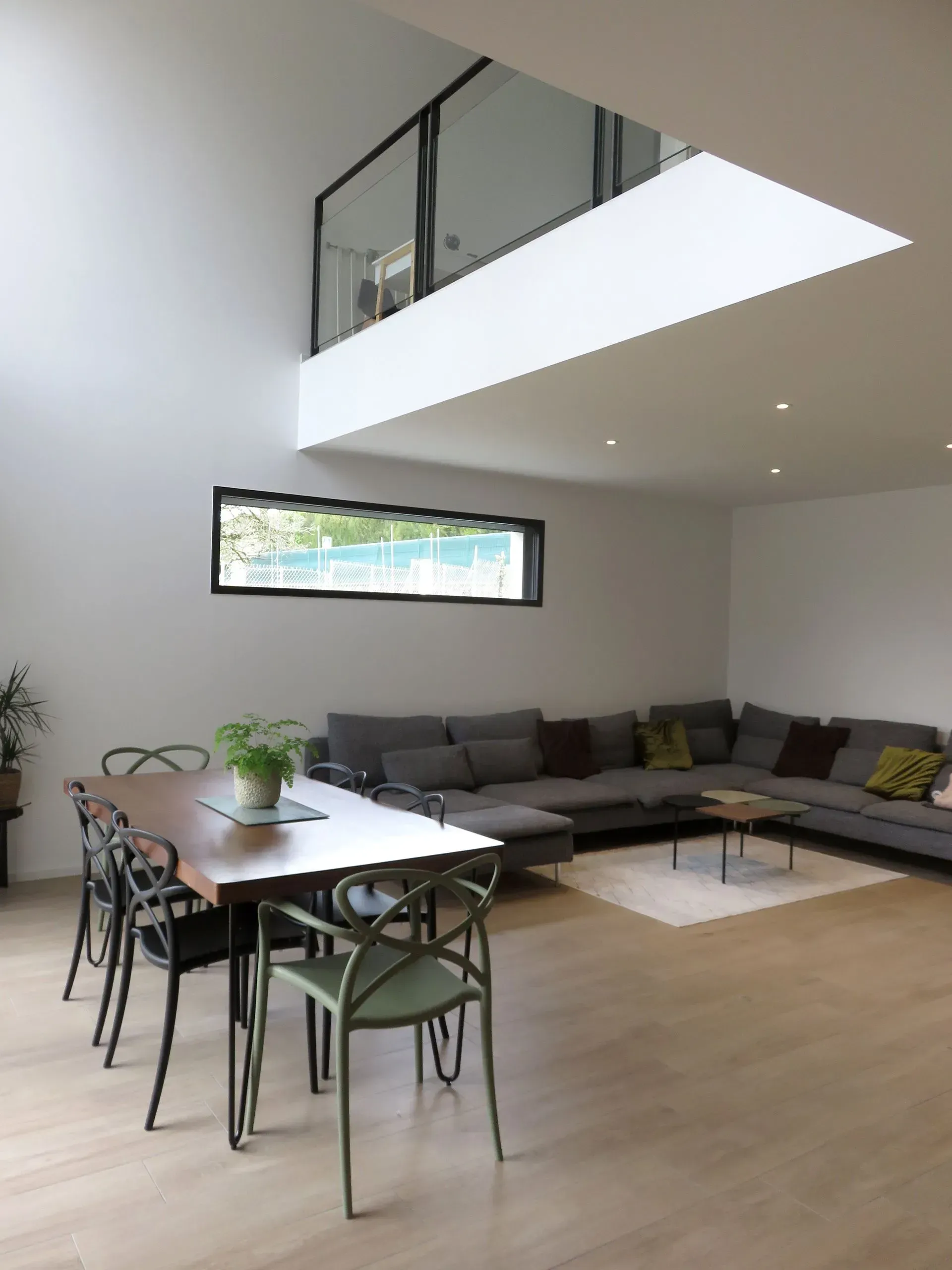 Un salon moderne et minimaliste avec un coin repas, une table en bois, un canapé d'angle gris et une mezzanine ouverte.