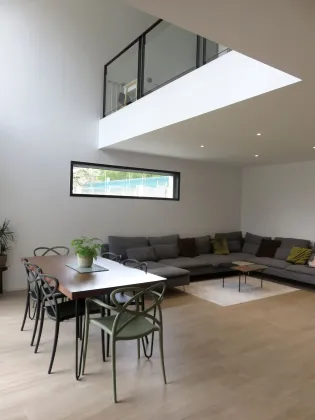 Un salon moderne et minimaliste avec un coin repas, une table en bois, un canapé d'angle gris et une mezzanine ouverte.